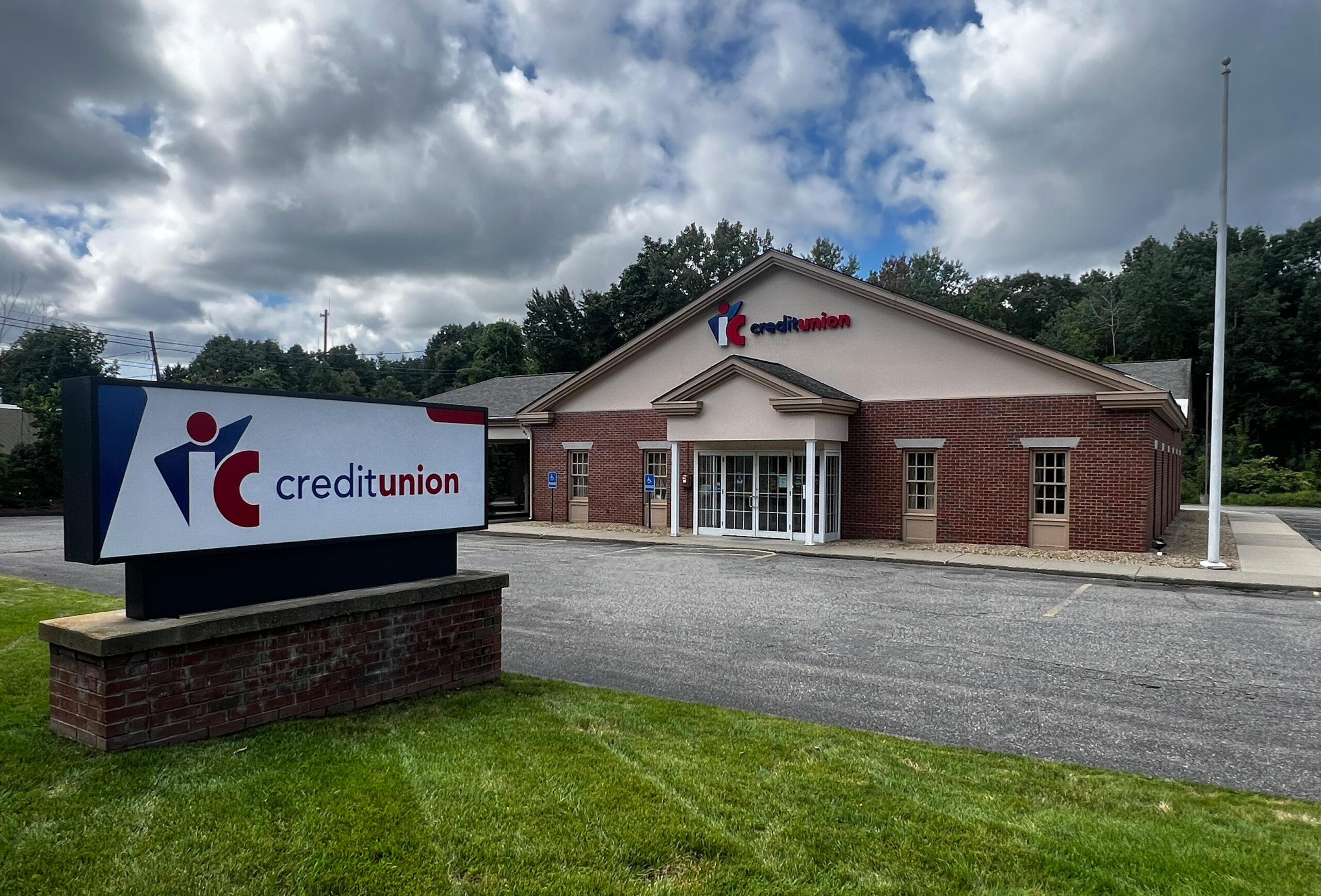 An image of the Ayer IC Credit Union exterior from the view of the front lawn. The building is made of brick and there are several windows. The image depicts two handicap parking spaces, and the weather is partly cloudy