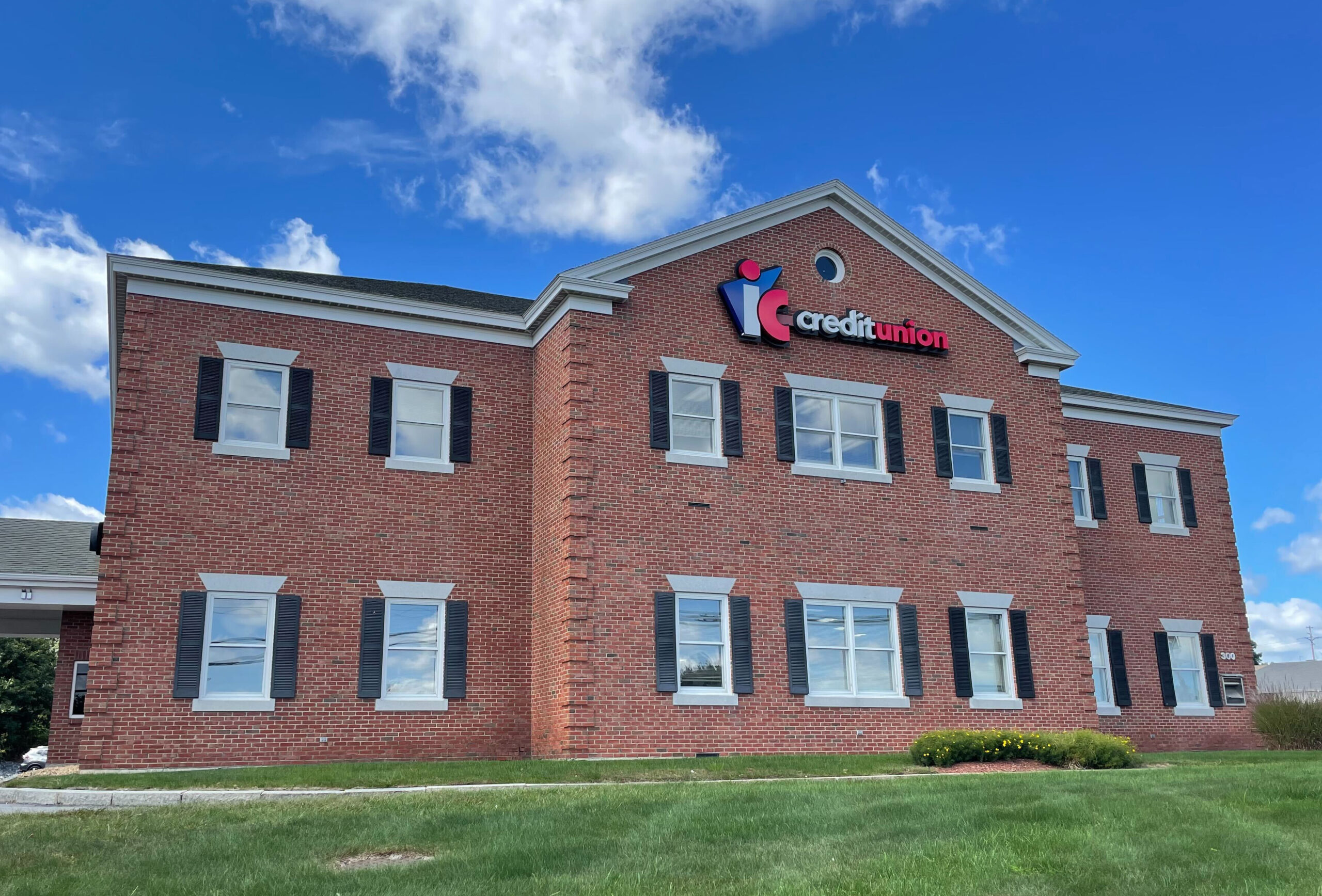 An image of the Fitchburg IC Credit Union main office from the view of the front lawn. The exterior of the building is brick, with several windows and black shutters. The sky is blue with a few clouds