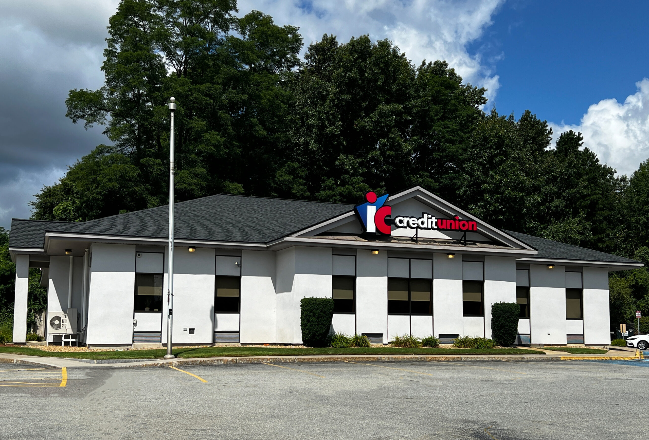 An image of the Leominster IC Credit Union Branch exterior from the view of the front parking lot. The 2 story building is white with several windows. There are full green trees behind the building, and the sky is blue with a few clouds