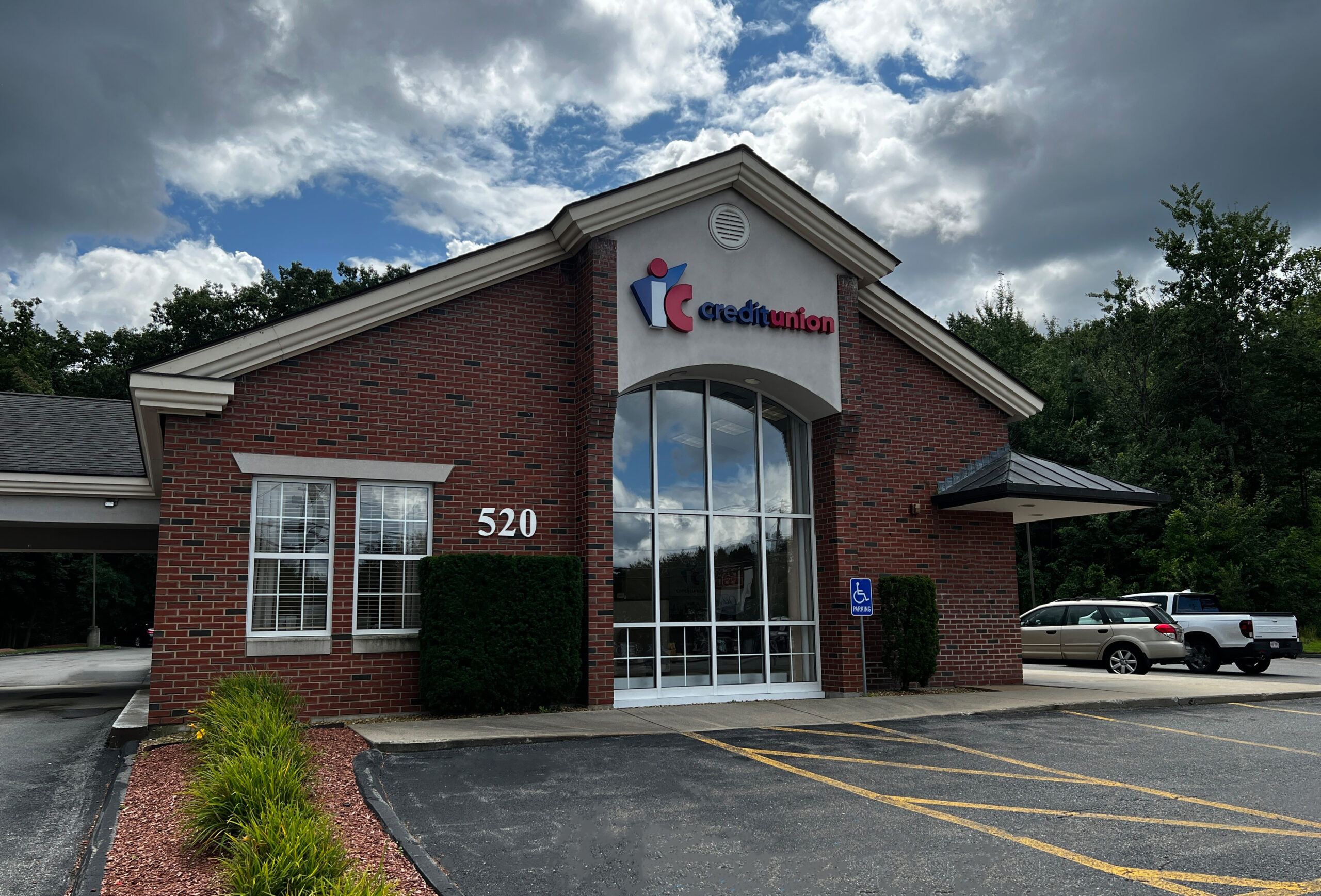 An image of the Fitchburg-Parkhill IC Credit Union exterior from the view of the front parking lot. The building is made of brick and has several large windows. There are cars parked outside the building, and the image captures an available handicap parking space. There is full greenery surrounding the building, and the sky is partly sunny