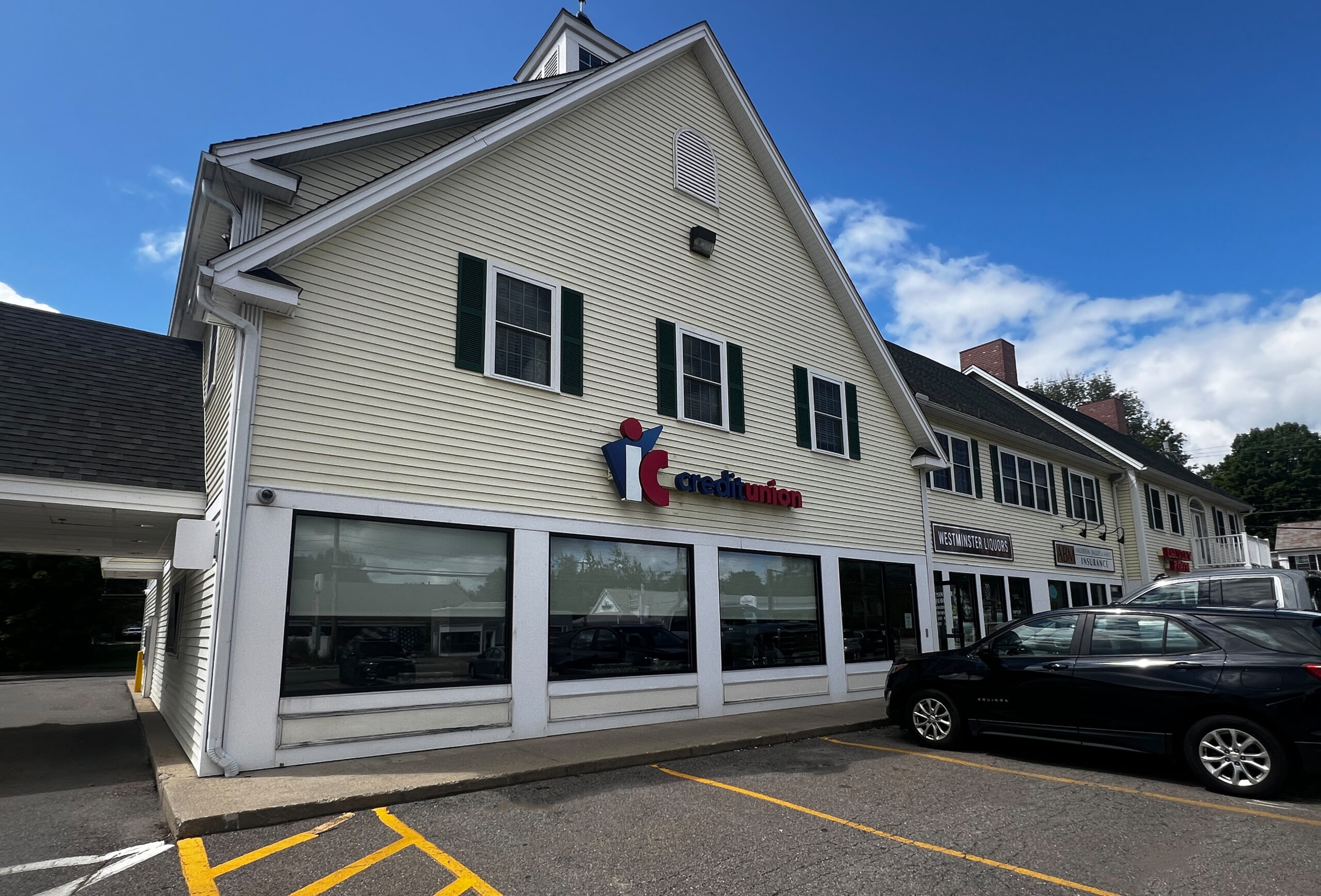 An image of the Westminster IC Credit Union exterior from the view of the front parking lot. The building has yellow siding, and a few windows with black shutters. The branch appears to be located in a plaza, and there are cars parked in the parking lot in front of it.