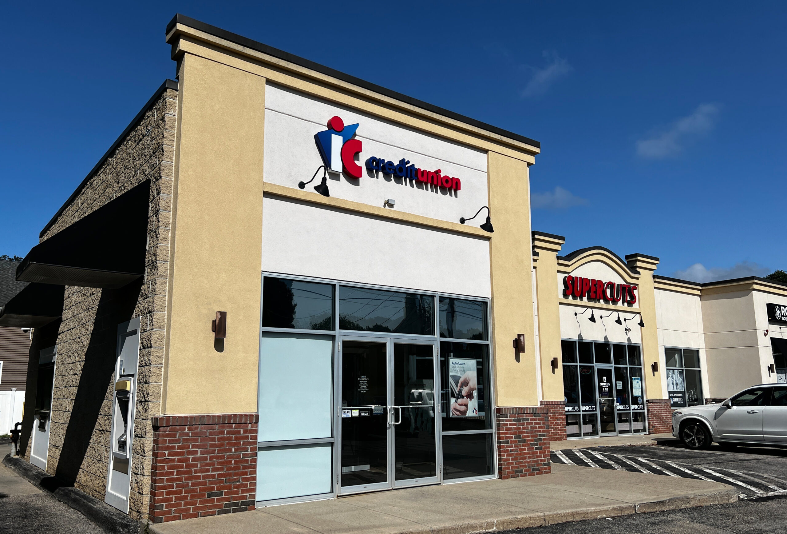 An image of the Worcester IC Credit Union Branch exterior from the view of the front parking lot. The building is light brown with brick accents and windows. The building appears to be in a plaza, and the walk-up ATM is visible. The sky is blue with a few clouds